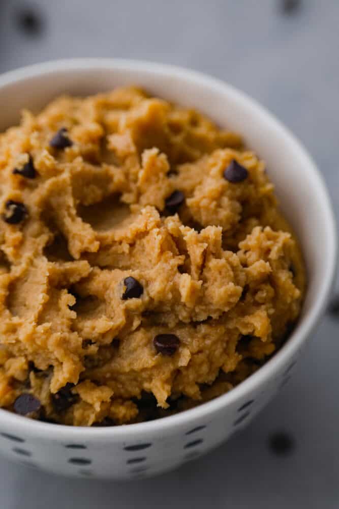 Who needs actual cookie dough when you can have chickpea cookie dough? It's the guilt-free treat that will have you feeling like a million bucks! Closeup of chocolate chip cookie dough in a white bowl.