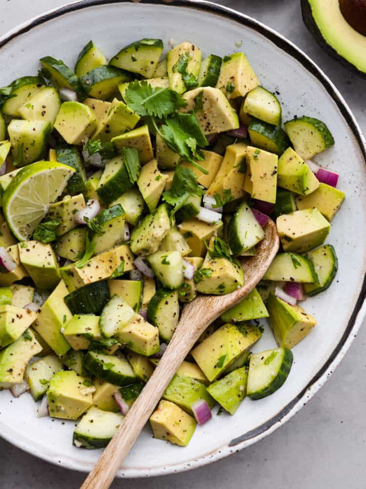 Cucumber avocado salad is the ultimate combination of creamy and crisp, tossed in a mouthwatering dressing made of lime juice, olive oil, and garlic! It's light, flavorful, and perfect for serving alongside all of your favorite summer meals. Cucumber avocado salad in a white bowl with a wooden spoon.